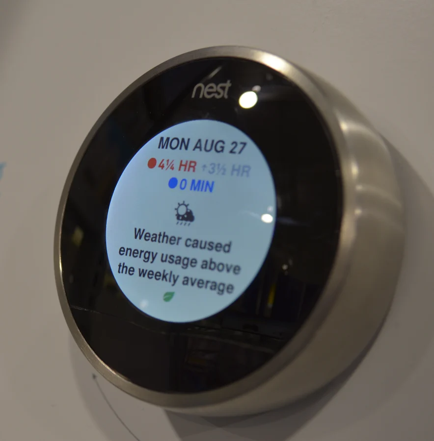 A Nest Learning Thermostat mounted on a wall, its round display showing a day of energy history and the line Weather caused energy usage above the weekly average.